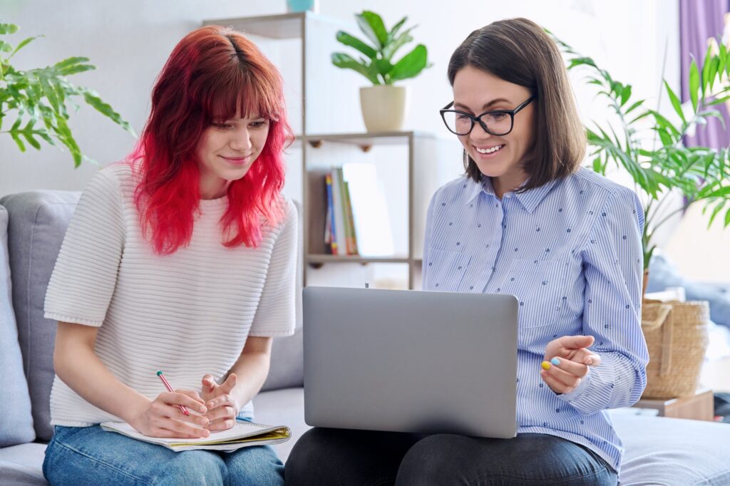 Female teacher teaches teenage girl, sitting together on couch in office, using laptop